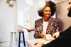 Smiling woman sitting at a table with coffee, smiling at someone.