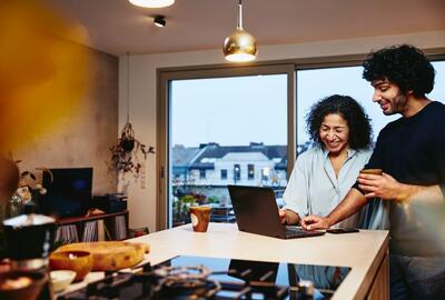 en leende kvinna och man står bredvid varandra vid köksbänken och tittar på en bärbar dator.