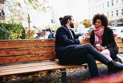 Man och kvinna med drinkar sitter på en bänk utomhus och samtalar.