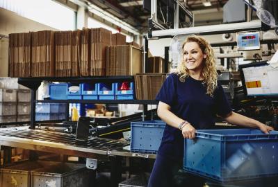 leende kvinna står bredvid ett transportband och packar ner produkter i en plastlåda