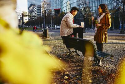 Man and woman holding drinks smiling and having a conversation. Man is sitting down on bench.