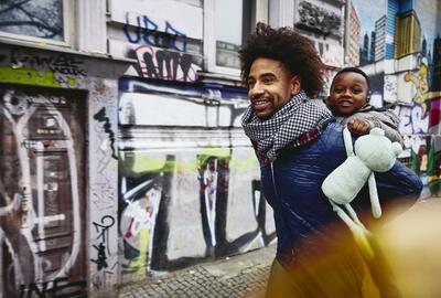 Smiling man with kid on his back walking through a city.