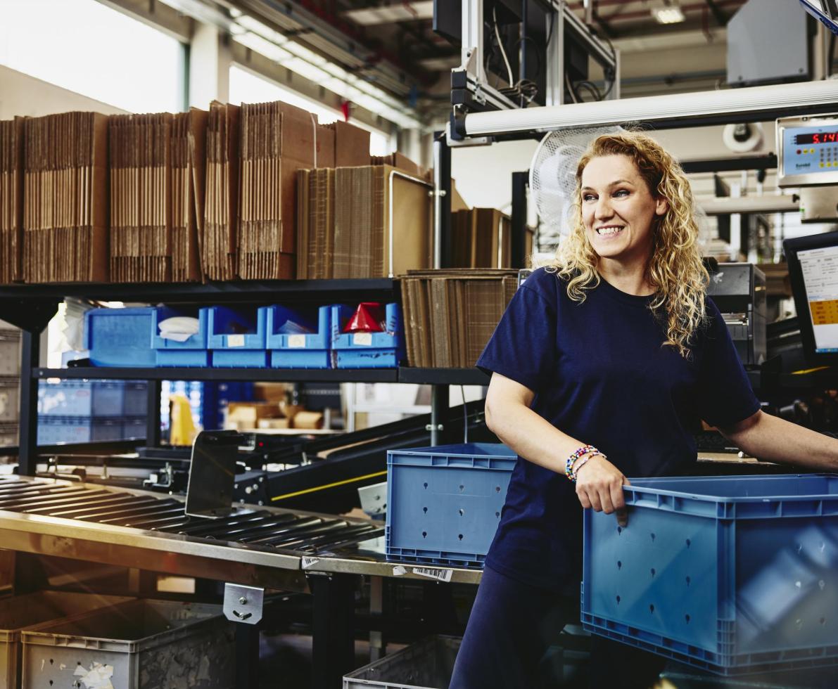 leende kvinna står bredvid ett transportband och packar ner produkter i en plastlåda