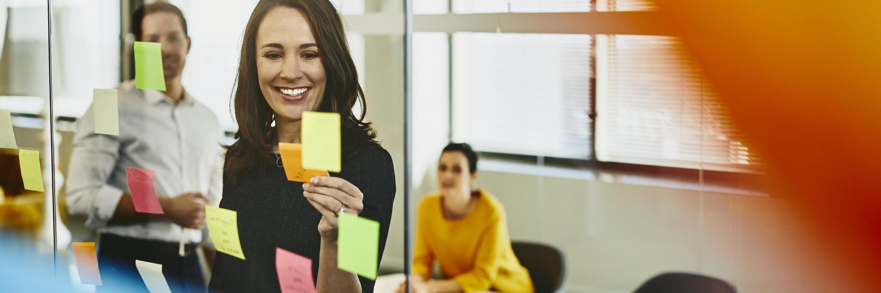 Two business women and a man in an office putting sticky notes on a window. Primary colors: yellow and blue.