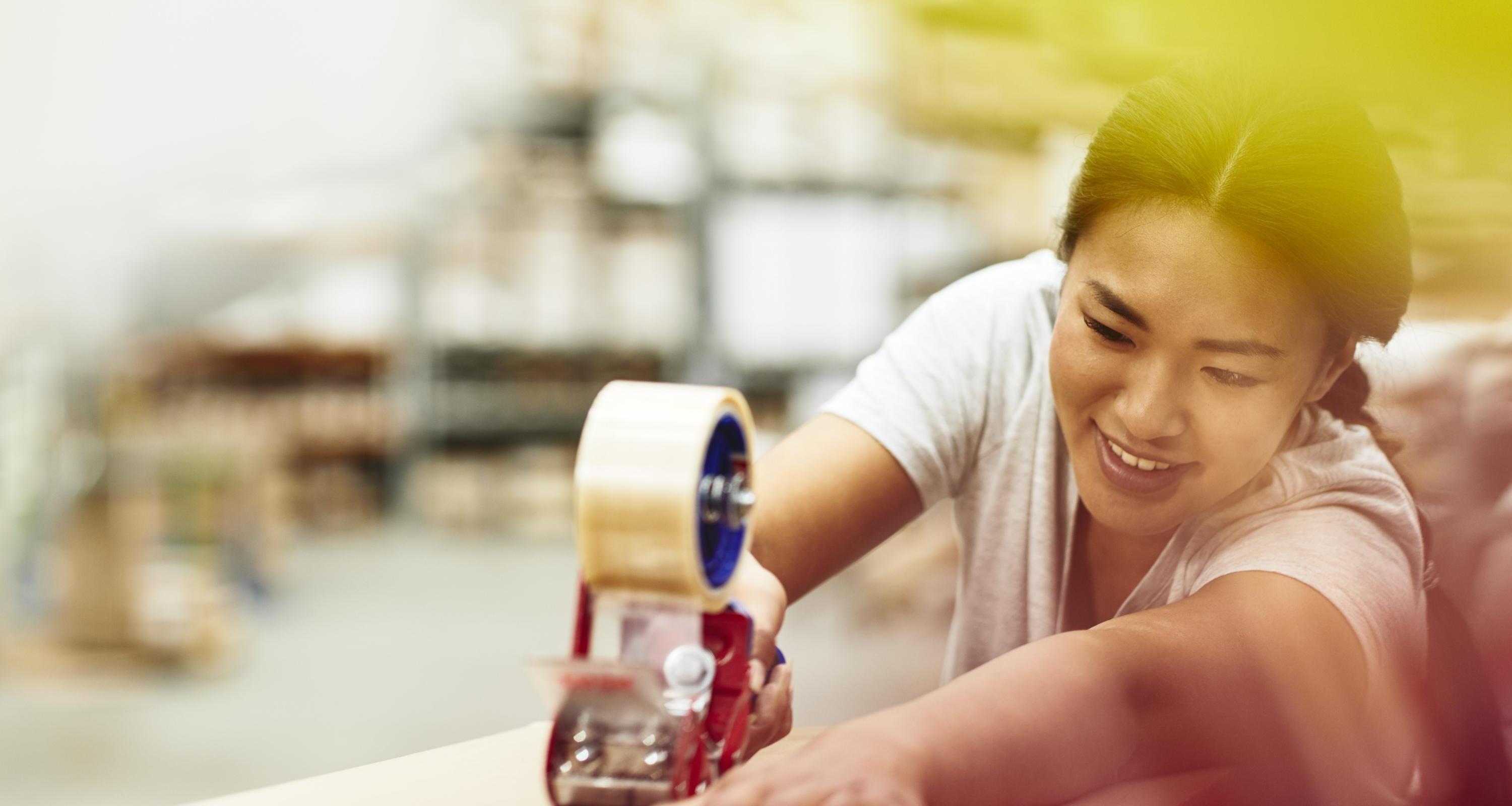 Female blue-collar worker enjoying taping a box. Logistics environment. Hair up. Braided. Asian woman. Primary color yellow.