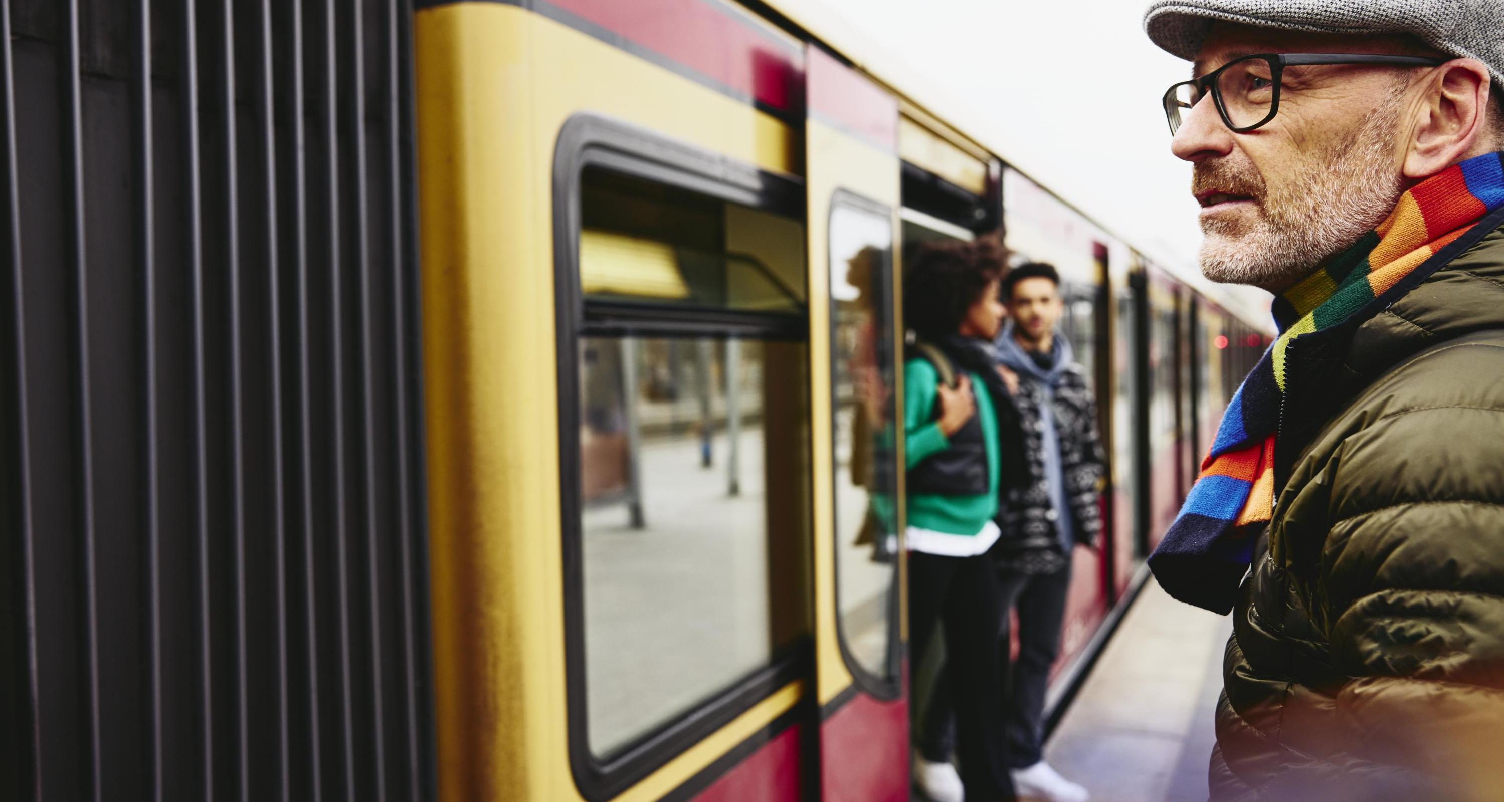 Man standing next to a train. Two people getting out of a train in the background.