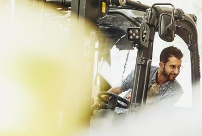 Male caucasian worker operating a forklift in a logistics environment. Looking to his left. Blue-collar. Smiling.  Wearing glasses. Groomed beard and moustache. Checkered shirt. Primary color cream.
