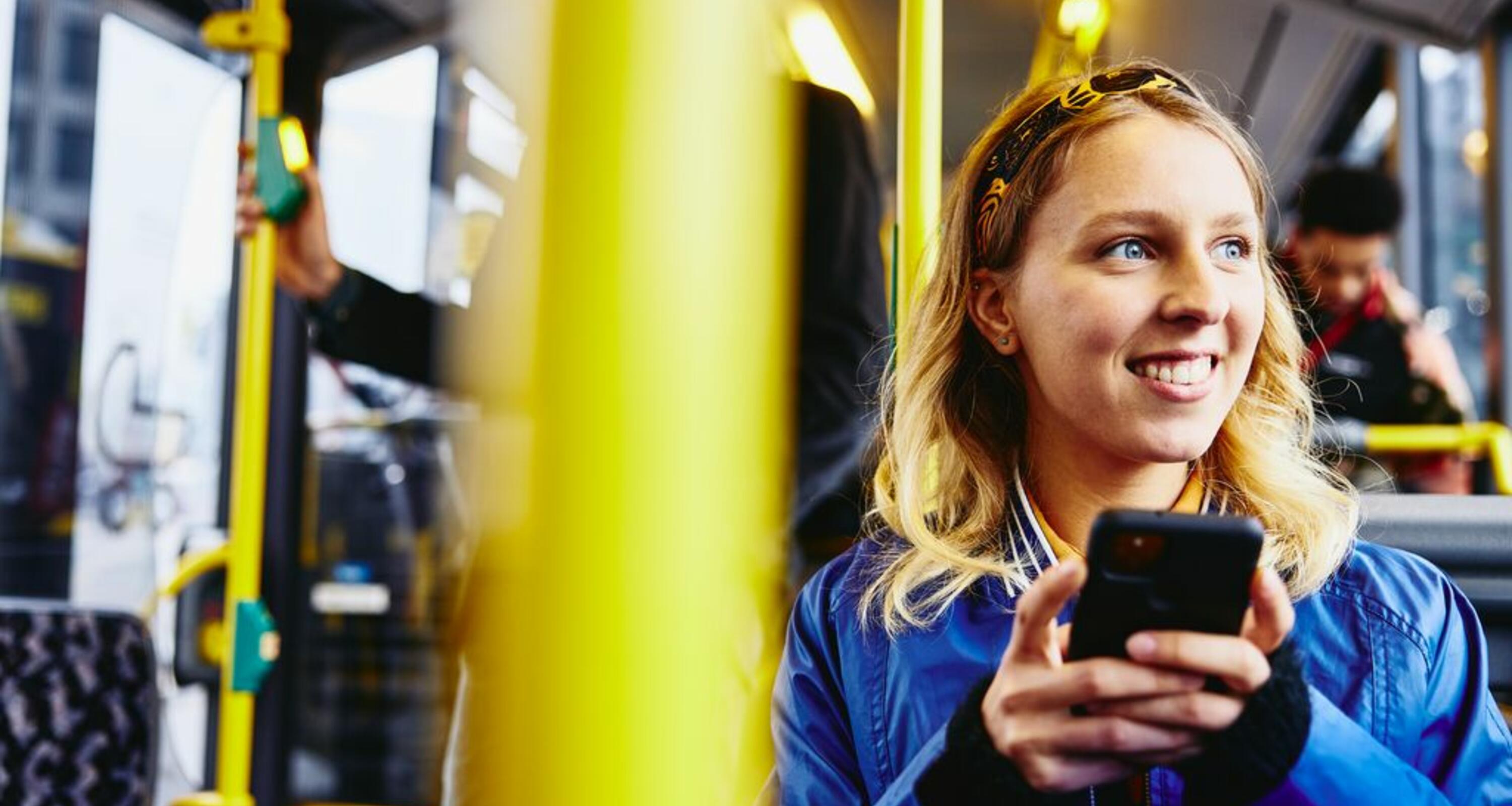 Leende kvinna sitter på bussen och tittar ut medan hon håller sin telefon i handen
